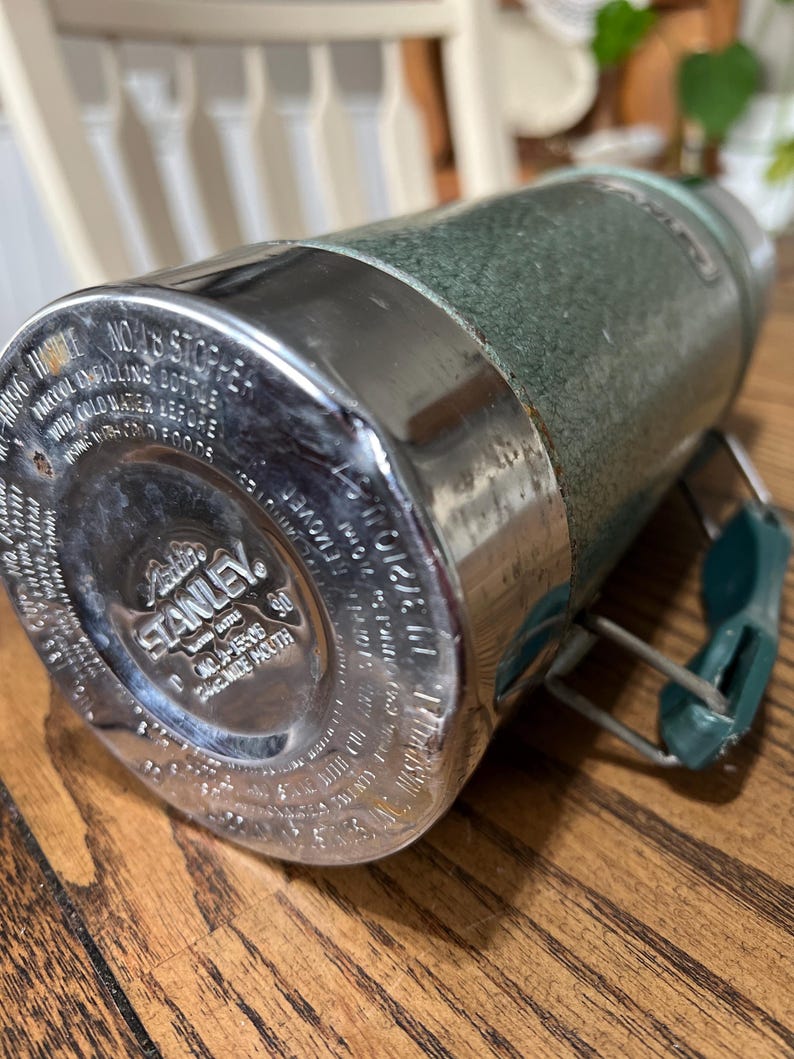 Puede incluir: Termo Stanley vintage, verde texturizado con detalles met&aacute;licos plateados. La base presenta el logo "Aladdin Stanley". El asa es verde. El termo est&aacute; sobre una superficie de madera. Dise&ntilde;ado para mantener l&iacute;quidos fr&iacute;os.