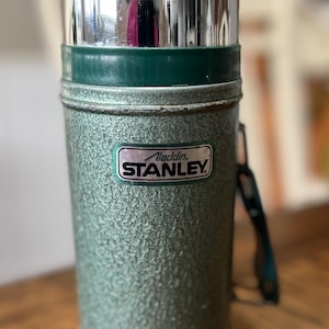 Puede incluir: Termo Aladdin Stanley verde vintage con acabado texturizado y tapa plateada brillante. El termo tiene un asa verde y una etiqueta que dice "Aladdin Stanley". Un recipiente aislado cl&aacute;sico para bebidas.