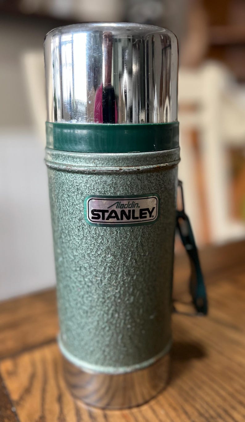 Puede incluir: Termo Aladdin Stanley verde vintage con acabado texturizado y tapa plateada brillante. El termo tiene un asa verde y una etiqueta que dice "Aladdin Stanley". Un recipiente aislado cl&aacute;sico para bebidas.