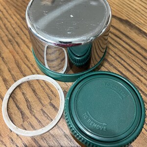 Puede incluir: Un recipiente de acero inoxidable brillante con base y tapa verdes, junto con un anillo de goma blanco. La tapa tiene las palabras "TO REMOVE" y "TO TIGHTEN" impresas. El recipiente est&aacute; dise&ntilde;ado para el almacenamiento.