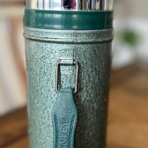 Puede incluir: Termo Stanley vintage en acabado verde texturizado con tapa plateada brillante. El termo tiene un asa de cuero verde con el logotipo de Stanley. Un art&iacute;culo cl&aacute;sico para mantener las bebidas calientes o fr&iacute;as.
