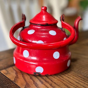 May include: A red enamel teapot with white polka dots. The teapot has a curved handle and a spout. The lid has a small knob. The teapot is sitting on a wooden surface.