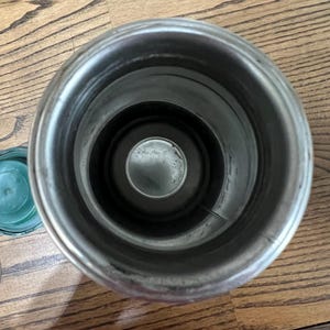 Puede incluir: Un recipiente aislado abierto de acero inoxidable con un interior oscuro. Una tapa verde y un anillo plateado son visibles a la izquierda. El recipiente est&aacute; sobre una superficie de madera.