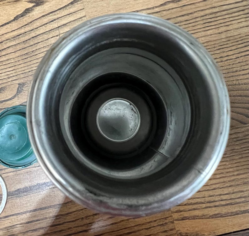 Puede incluir: Un recipiente aislado abierto de acero inoxidable con un interior oscuro. Una tapa verde y un anillo plateado son visibles a la izquierda. El recipiente est&aacute; sobre una superficie de madera.