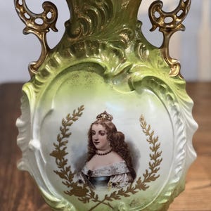 May include: An antique porcelain vase with a portrait of a woman in a crown, framed by gold laurel leaves. The vase features ornate gold handles and accents, with a green and white color scheme. A decorative piece.