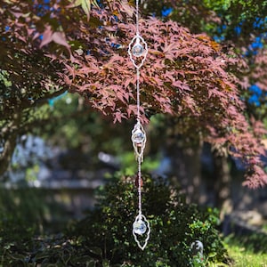 Puede incluir: Un adorno colgante vertical que presenta tres marcos de metal plateado, cada uno con forma ovalada puntiaguda y bordes festoneados. Una esfera de cristal transparente y facetado está suspendida en el centro de cada uno de los tres marcos.