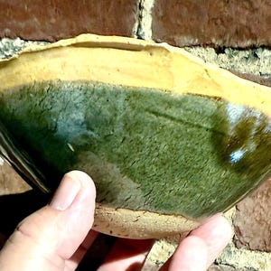 May include: A handmade ceramic bowl with a unique design. The bowl features a two-tone glaze, with a green interior and a yellow-brown rim. The bowl is held in a hand, showcasing its shape and texture.