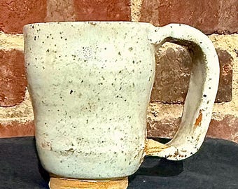 May include: A handmade ceramic mug with a speckled, off-white glaze. The mug has a large, curved handle and a slightly flared base. The base is a light tan color. The mug is set against a brick wall.