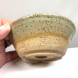 May include: A speckled ceramic bonsai pot with a rounded shape. The pot features a light green glaze on the top half, transitioning to a tan base. A small, square stamp is visible on the side.