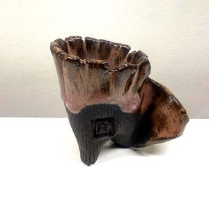 May include: A handcrafted ceramic planter with a unique, organic design. The planter features a dark brown base, transitioning to a lighter brown and pinkish rim. The textured surface gives it a rustic, natural appearance. The planter is suitable for small plants.