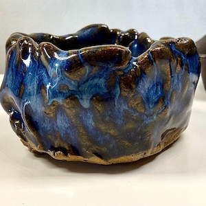 May include: A handcrafted ceramic bowl with a unique, organic shape. The bowl features a glossy, blue glaze that drips down the sides, contrasting with the brown, textured rim. The interior is a deep blue, creating a striking visual effect.