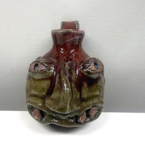 May include: A handmade ceramic bottle with a face design. The bottle features a dark red top and a green and brown face with protruding eyes and teeth. The bottle has a rustic, textured appearance and is likely a decorative piece.