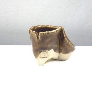May include: A handmade ceramic cup with a unique, asymmetrical design. The cup features a two-tone color scheme, with a light beige base and a dark brown upper portion. The cup has a textured surface and an embossed letter "R" on the base.
