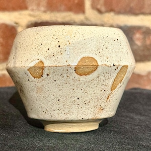 May include: A handmade, off-white ceramic bowl with a unique geometric shape. The bowl features a speckled texture with brown spots and accents. The design includes two large, circular brown spots and a long, thin brown streak.