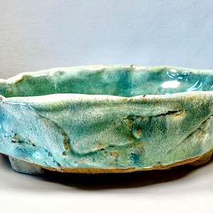 May include: A handcrafted ceramic bowl with a textured, irregular rim. The bowl features a blend of turquoise and green glazes, with hints of yellow and brown. The organic shape and color palette suggest a natural, earthy aesthetic. This decorative bowl is suitable for home decor.