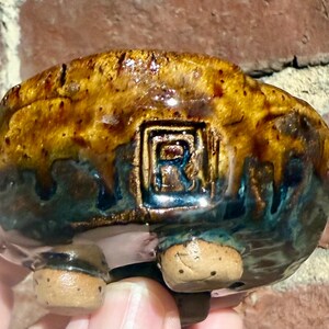 May include: A small, handcrafted ceramic bowl with a unique glaze. The bowl features a blend of brown, gold, and teal colors. The base has three small feet. The letter "R" is imprinted on the side of the bowl.