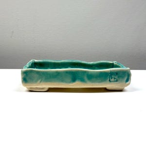 Może przedstawiać: Prostokątna ceramiczna donica z turkusową glazurą. Donica ma falisty górny brzeg i stoi na czterech małych nóżkach. Podstawa ma naturalny, nieszkliwiony kolor. Odpowiednia dla małych roślin lub sukulentów.