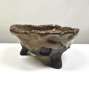 Puede incluir: Un cuenco de cerámica hecho a mano con un borde texturizado e irregular y tres patas cortas. El cuenco tiene un esmalte marrón y beige con detalles en marrón oscuro. La base es de color marrón oscuro.