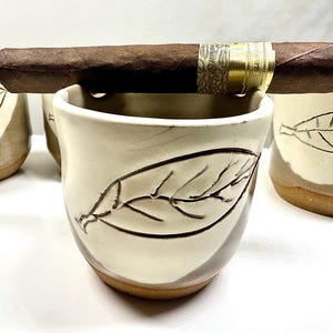 May include: A handmade ceramic cigar ashtray with a leaf design. The ashtray is off-white with brown accents and a brown base. A dark brown cigar rests on the rim of the ashtray. The ashtray is designed to hold a cigar.