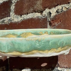 May include: A handmade ceramic bowl with a rectangular shape. The bowl features a light green glaze with a yellow and white accent. The bowl is held by a hand in front of a brick wall.