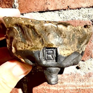 May include: A small, handmade ceramic bowl with a unique, textured surface. The bowl features a two-tone color scheme, with a dark brown top and a gray base. The bowl has a small, square design on the side and is supported by three small feet.