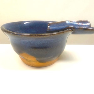 May include: A handmade ceramic bowl with a built-in rest for a cigar or cigarette. The bowl is a deep blue with a brown rim and a golden-brown base. The ashtray is a unique piece of pottery.