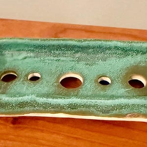 May include: A rectangular, handmade ceramic soap dish with a textured, turquoise-green glaze. The dish has five circular drainage holes and a cream-colored base. The soap dish is a decorative and functional bathroom accessory.