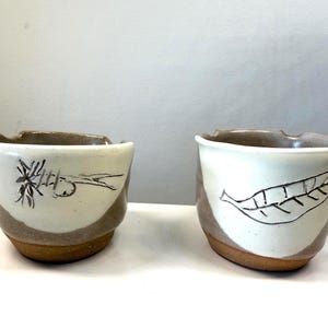 May include: Two handmade ceramic bowls. Each bowl has a white upper section with a hand-drawn black design, one with a plant and the other with a leaf. The lower section and base are brown.