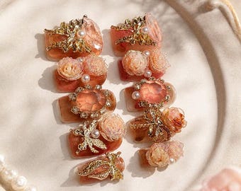 May include: A collection of ten ornate, artificial nails. Each nail features a base of varying shades of peach and brown, adorned with gold-colored embellishments, pearls, and miniature floral designs. Some nails have large, faceted gems.