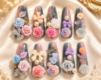 May include: A collection of ten long, artificial nails with a marbled gray and pink base. Each nail is adorned with 3D floral designs, bows, pearls, and decorative accents in various colors, including blue, pink, and white. The nails are arranged on a satin surface.