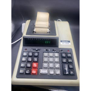 May include: A beige Unisonic XL-131X printing calculator with a black control panel, a digital display showing 0.00, and a roll of paper tape. The device features a numeric keypad with gray keys, black function keys, and red clear buttons.