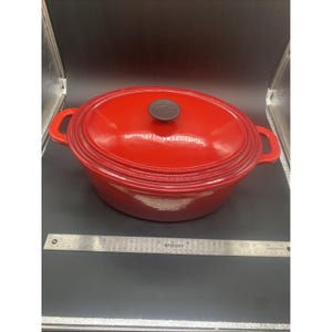 May include: A bright red oval enameled cast iron Dutch oven with two side handles and a matching lid featuring a black knob with the text Cook's Essentials. A silver metal ruler is placed in front for scale reference.