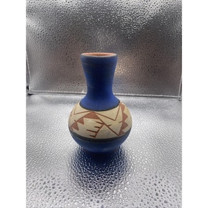 May include: A small, blue ceramic vase with a narrow neck and a rounded body. The vase features a geometric pattern in brown and cream colors around the middle. The rim is a reddish-brown color.