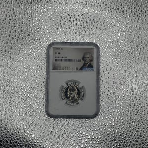 Può includere: Una moneta d'argento racchiusa in un supporto di plastica trasparente. La moneta presenta il profilo di un uomo ed è contrassegnata con "1963 SC PF 68". Il supporto mostra anche un ritratto e il logo NGC.