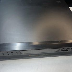 Puede incluir: Un cambiador de CD negro con capacidad para 5 CD. El dispositivo tiene un diseño rectangular con botones y una pantalla. El texto "5 CD CHANGER Disc Ex-change System" es visible en la parte frontal.