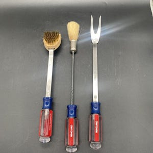 May include: Three Craftsman tools with clear and red handles. The left tool has a brass wire brush head. The center tool features a round, light-colored bristle brush. The right tool is a two-pronged metal fork. Each handle is marked Craftsman.