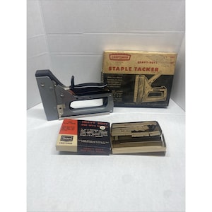Puede incluir: Una grapadora resistente Craftsman gris y negra con su caja de cartón original con texto en rojo y negro. Al lado hay una caja abierta de grapas de alambre .050 con una etiqueta roja, blanca y negra y la marca Sears.