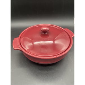 May include: A round, deep red ceramic casserole dish with a matching lid. The dish features two integrated side handles and a central knob on the lid. The surface has a smooth, glossy finish, reflecting light across the curved, circular form.