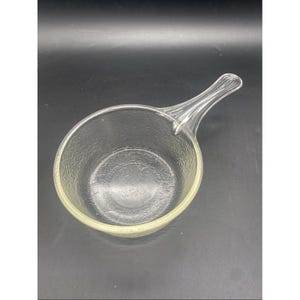 May include: A clear glass saucepan with a round bowl and a single handle. The exterior of the bowl features a textured, pebbled surface pattern, while the handle has a smooth, ribbed design. The glass is transparent and reflects light.