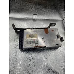 May include: A silver car stereo component with a black front and a metal bracket. The component has several labels with text and numbers. The bracket is brown and attached to the top of the component.