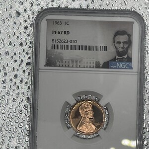 Può includere: Una moneta da 1 centesimo Lincoln 1C del 1963, classificata PF 67 RD, racchiusa in un supporto protettivo trasparente. La moneta presenta un ritratto di Abraham Lincoln e il testo "LIBERTY" e "1963". Il supporto include la certificazione NGC e il numero di serie.