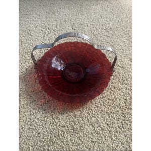 Könnte beinhalten: Eine runde, tiefrote Glasschale mit einem strukturierten, gewellten Rand und einer glatten Mitte. Die Schale ist mit einem silberfarbenen Metallgriff ausgestattet, der ein dezentes, sich wiederholendes Fischgrätenmuster aufweist.
