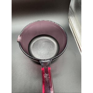 May include: A purple glass saucepan with a black interior and a red handle. The saucepan has a pouring spout and a handle with text. The handle is red and the pan is purple.