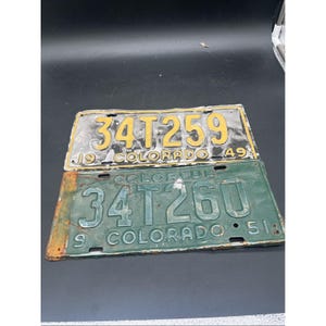 Puede incluir: Dos placas de matrícula de metal vintage de Colorado. La placa superior es negra con texto amarillo que dice 34T259 y 19 49. La placa inferior es verde con texto en relieve que dice COLORFUL 34T260 y 19 51, con óxido visible en el borde izquierdo.