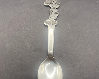 Op de afbeelding: Een zilverkleurige metalen lepel met een gebeeldhouwde figuur van Mickey Mouse aan de bovenkant van de steel. De lepel heeft een gladde, gepolijste kom en een slanke steel die aansluit op het gedetailleerde karakterontwerp aan het uiteinde.