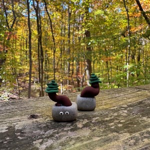 May include: Two whimsical clay sculptures resembling small trees, each with a brown trunk and a green, layered top. They are set in gray, stone-like pots with googly eyes, placed on a weathered wooden surface against a backdrop of autumn foliage.