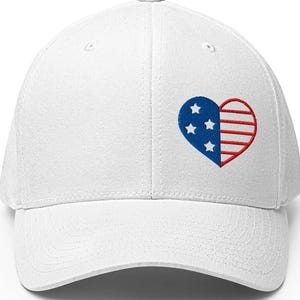 Könnte beinhalten: Weiße Baseballkappe mit einem gestickten Herzdesign. Das Herz ist in zwei Bereiche unterteilt: einen mit blauen Sternen und einen mit rot-weißen Streifen, die ein patriotisches Thema hervorrufen. Die Kappe hat einen gebogenen Schirm und eine strukturierte Passform.