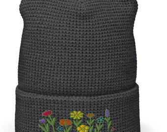 Puede incluir: Un gorro gris oscuro con un borde doblado. El borde presenta un diseño floral bordado con flores rojas, naranjas, amarillas, azules y moradas. El gorro está hecho de un material de punto texturizado.