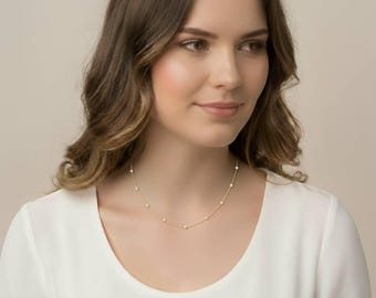 May include: A delicate gold necklace featuring small, evenly spaced pearls. The necklace is worn by a person wearing a white top. The pearls are round and white, contrasting with the gold chain. The necklace is a simple yet elegant piece of jewelry.
