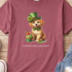 May include: Maroon t-shirt with a cartoon puppy wearing a green leprechaun hat and bow tie. A cupcake with a strawberry is next to the puppy. The text "Cuteness is My Lucky Charm" is below the image.